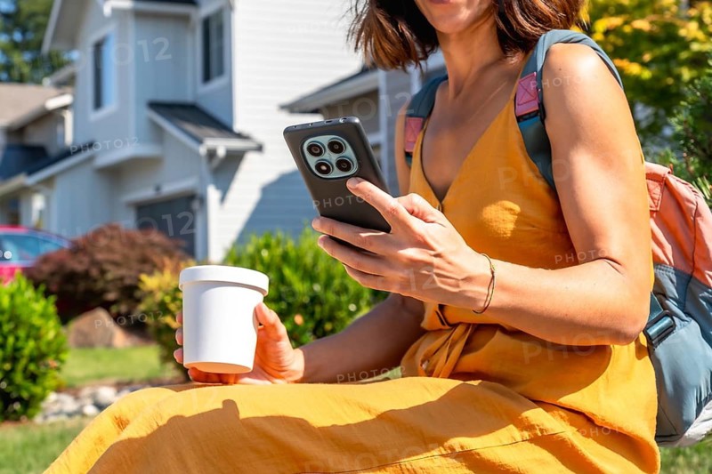 Woman in a yellow dress and backpack interacting with a smartphone while holding a coffee cup. Sitting outdoors in a sunny residential neighborhood. Representing modern urban lifestyle and technology