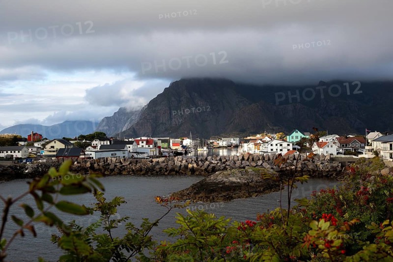 View of the picturesque fishing village against a gigantic mountain backdrop with deep clouds and autumn vegetation in the foreground, Henningsvær, Norway