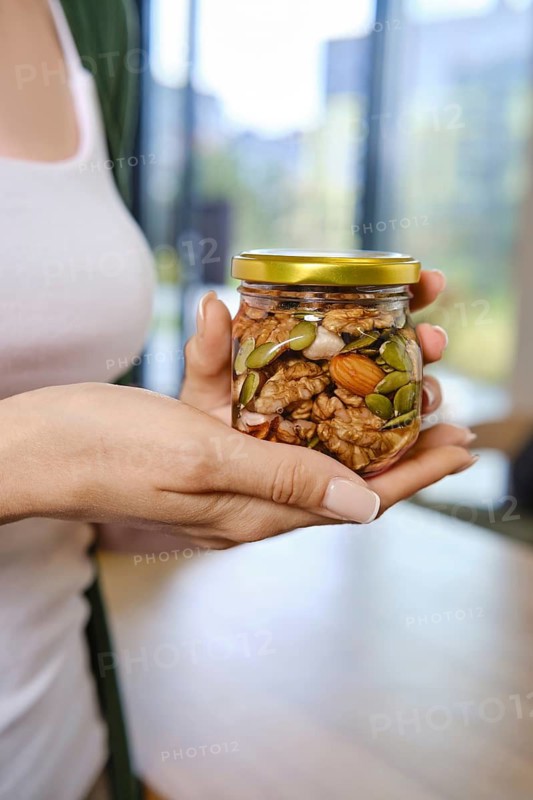 Person stands indoors in bright room with large windows holding a jar of honey filled with mixed nuts and seeds. Sunlight enters illuminating the contents of jar