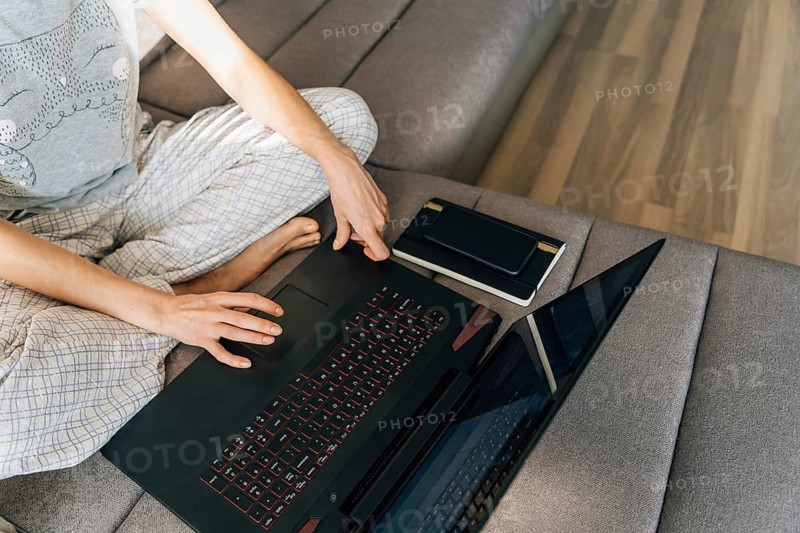 A young woman in pajamas sits on the couch and uses a laptop, Person sitting cross-legged on a couch using a laptop with a notebook nearby, in a casual and relaxed home setting