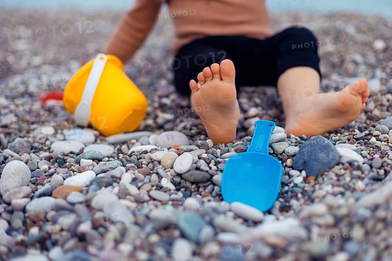 Two years girl toddler sits on a pebbly beach, playing with a yellow bucket and blue shovel. Sunlight shines on the childs bare feet and the colorful toys while waves gently lap in the background