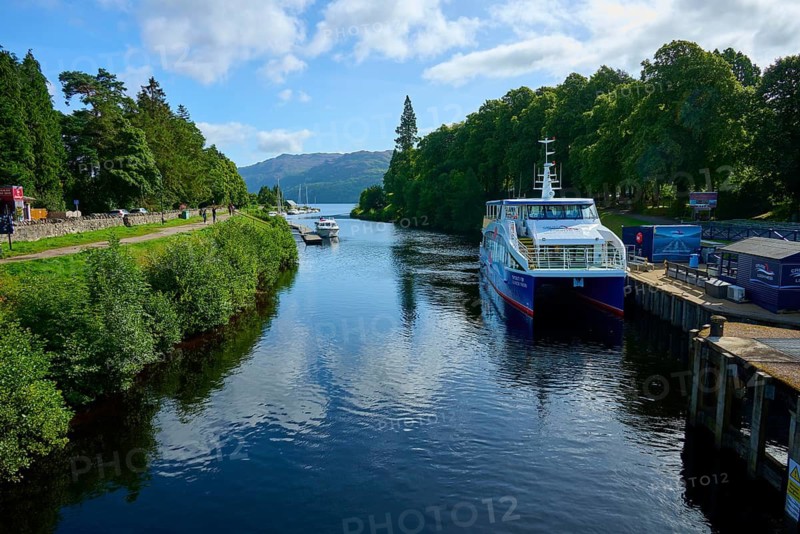 A large boat sails on a quiet river surrounded by trees and rolling hills, Caledonian Canal, Fort Augustus, Loch Ness, Scotland, United Kingdom, Great Britain