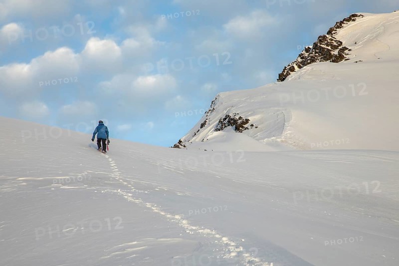 A skier in a blue jacket ascends a pristine snowy mountain slope, leaving tracks beneath a partly cloudy sky, capturing the essence of winter exploration and adventure