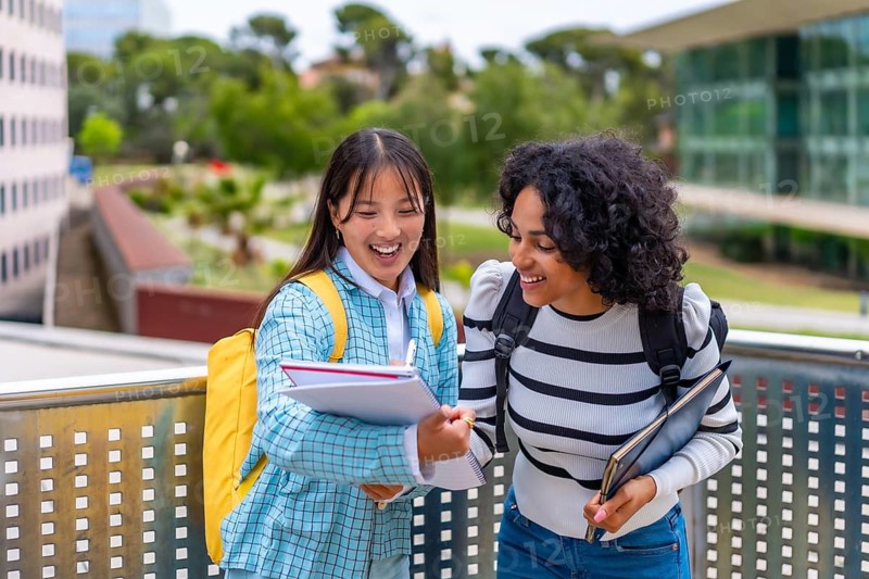 Two smiling multi-ethnic female students having fun while reading notes in the university campus