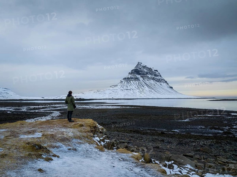 A lone traveler stands near the snow-dusted Kirkjufell mountain and its reflection in the calm waters, capturing the serene and majestic Icelandic winter landscape
