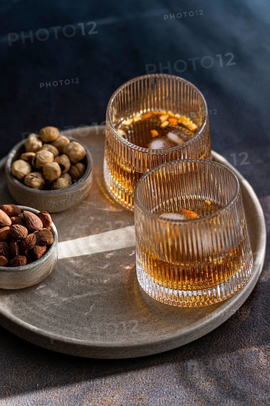 Elegantly presented cognac-based cocktails with cinnamon and star anise, accompanied by bowls of almonds, peanuts, and hazelnuts, stylishly served on a textured tray under soft lighting