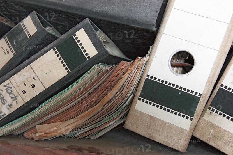 Office scene around 1982, lost place, abandoned location, classic file folders on a wooden shelf, some of the folders are labelled in detail by hand and provide information about their contents, mixed up, dirty