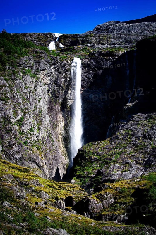Tall waterfall plunging into a rocky gorge surrounded by green vegetation and blue sky, view of a waterfall on a steep mountain wall with the Myklebustbreen glacier at the top, Vestland, Norway, Scandinavia