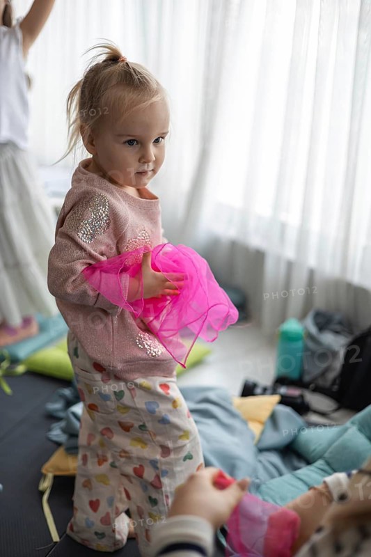 A young Caucasian girl with blonde hair holds a pink fabric while standing in a cozy home environment. The room is filled with soft cushions and natural light