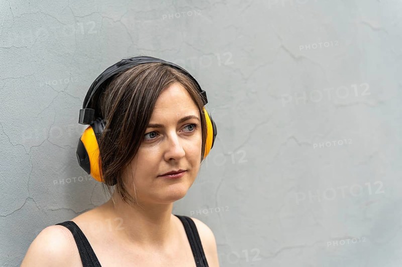 Close-up portrait of blonde woman 40s in yellow headphones against light wall, A woman wearing yellow and black headphones stands against a textured concrete wall, looking thoughtful