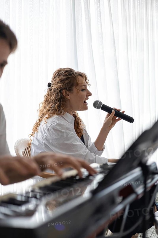 A young Caucasian woman with curly hair sings into a microphone. A hand plays a keyboard in the foreground. Bright, airy home setting with white curtains