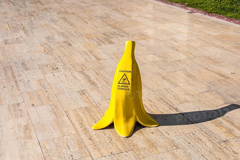 A yellow banana-shaped caution sign stands by a swimming pool. The pool is surrounded by modern buildings and palm trees, indicating a tropical setting