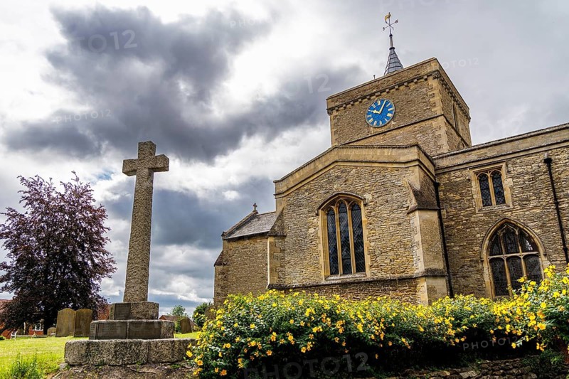 Stone war memorial cross and blooming yellow shrubs before St James the Great church tower, Bierton, Buckinghamshire, United Kingdom