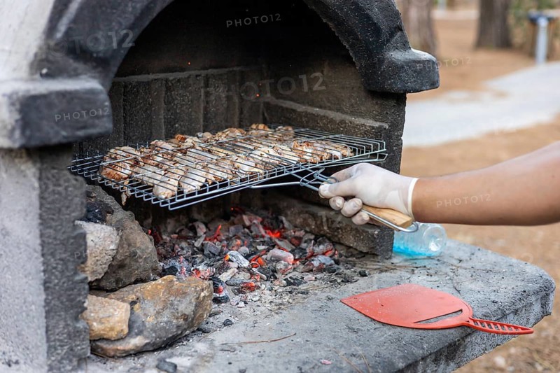 A hand with a glove holds a grill rack with cooked meat over hot coals. The scene is set in a park with trees in the background