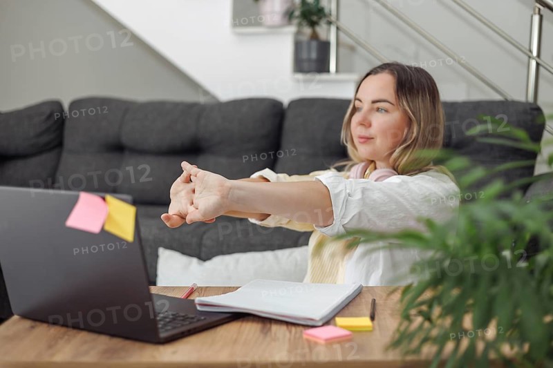 Young blond European woman with headphones sitting at wooden table with laptop and notebook, studying or working indoors, casual home environment