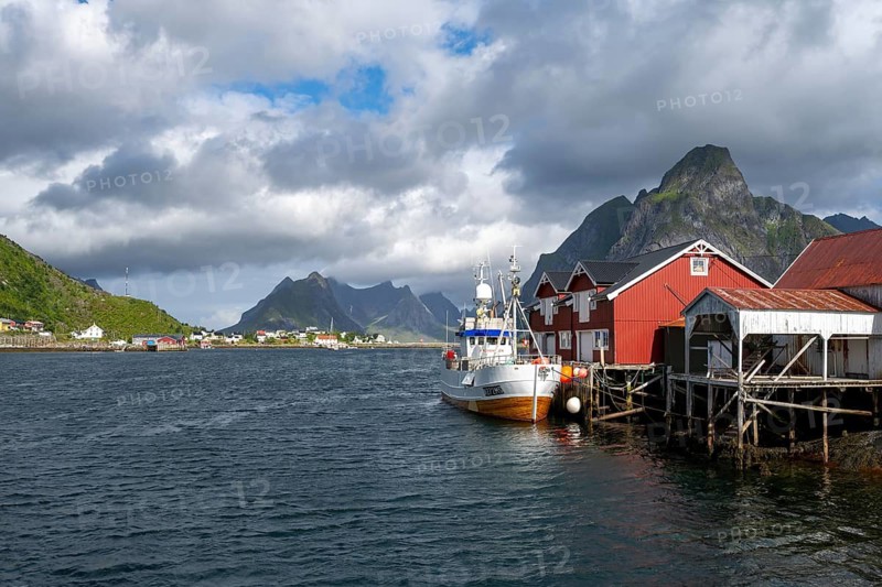 Ship and rorbuer, typical red wooden houses, stilt houses on the banks of Reinefjord, Reine, Lofoten, Norway