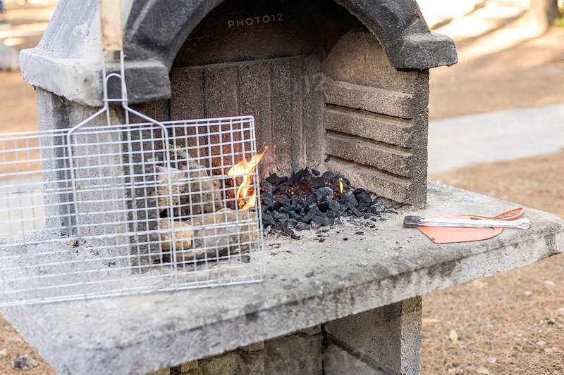 A stone barbecue grill with burning charcoal and flames. A metal grill rack is placed on top. The setting is outdoors with a natural background
