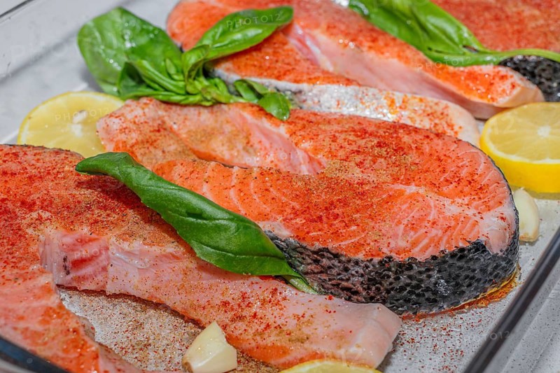 Fresh salmon fillets arranged in a glass dish with lemon slices and basil leaves. The fish is raw and ready for cooking. Healthy food and diet concept