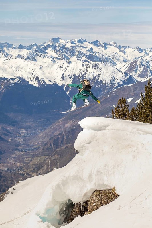 A snowboarder performs an exhilarating jump off a snow-covered ridge, surrounded by majestic mountain peaks. The scene captures the thrill and beauty of winter freeride adventures