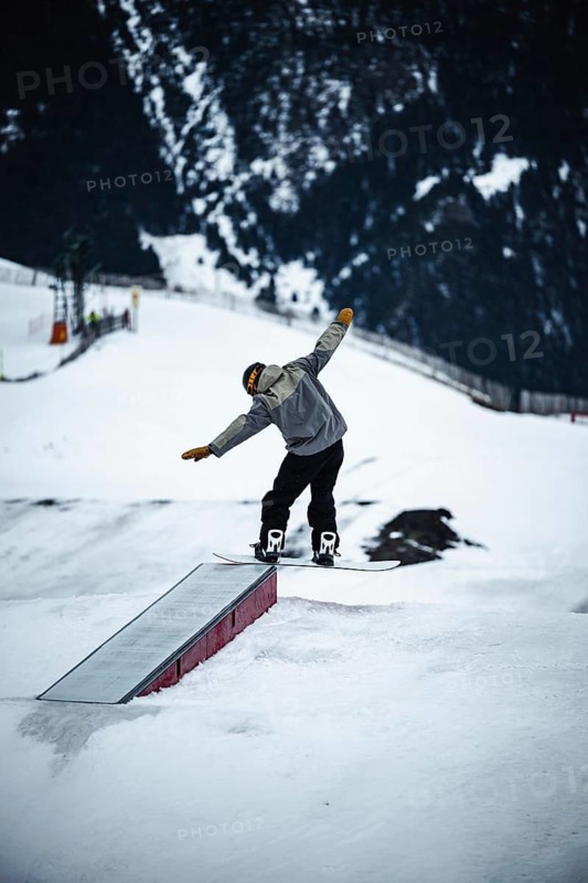 Snowboarder in motion, performing a trick on a snowy slope with mountainous backdrop in Andorra. Captures dynamic winter sports action, emphasizing adventure and skillful movement