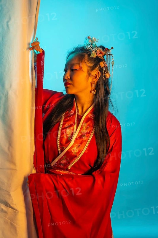 A woman in a red dress with a flowery headband. She is standing in front of a white curtain