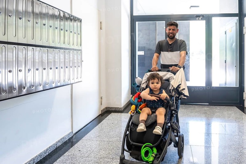 Father returning home, pushing a baby stroller with his young son in a residential building hallway, passing by mailboxes and enjoying family lifestyle together