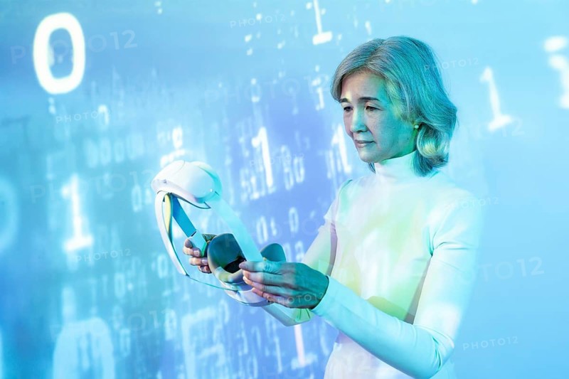 An elderly Asian woman engages with virtual reality technology, represented by a headset, set against a futuristic backdrop with digital coding and abstract blue hues