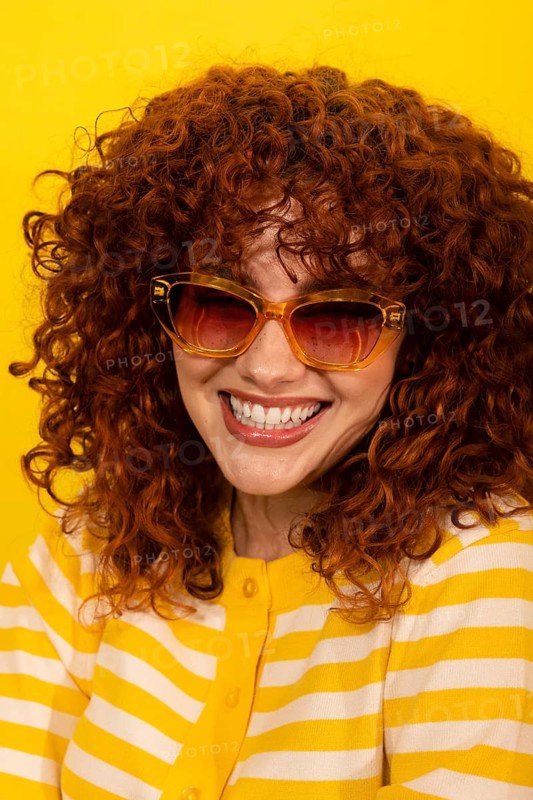 A woman with vibrant curly hair smiles confidently, wearing fashionable sunglasses and a yellow-striped outfit, set against a striking yellow background for a cheerful look