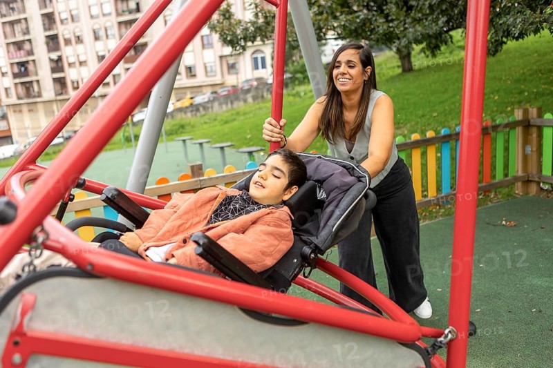 A mother joyfully pushes her child with special needs on an adaptive swing in a playground, surrounded by greenery and colorful play equipment, enriching their day