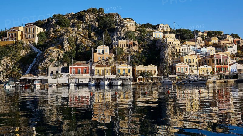 Houses on a rocky slope are reflected in the calm harbour water, boats are moored, Symi Town, Symi, Dodecanese, Greek Islands, Greece