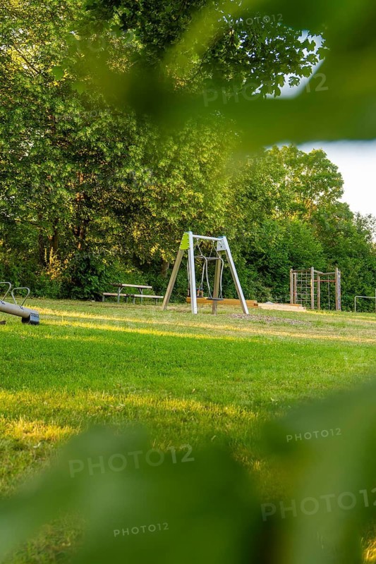 Playground on an open green field, surrounded by trees, partly covered by leaves in the foreground, playgrounds in Gechingen, Calw district, Germany