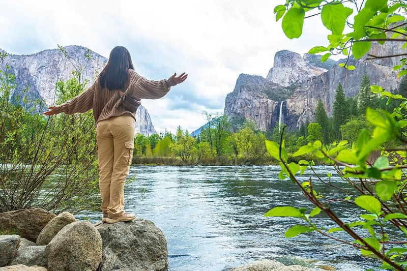 Woman standing on rocks by the merced river, spreading her arms wide, enjoying the vast scenery of yosemite national park with bridalveil fall and towering granite cliffs