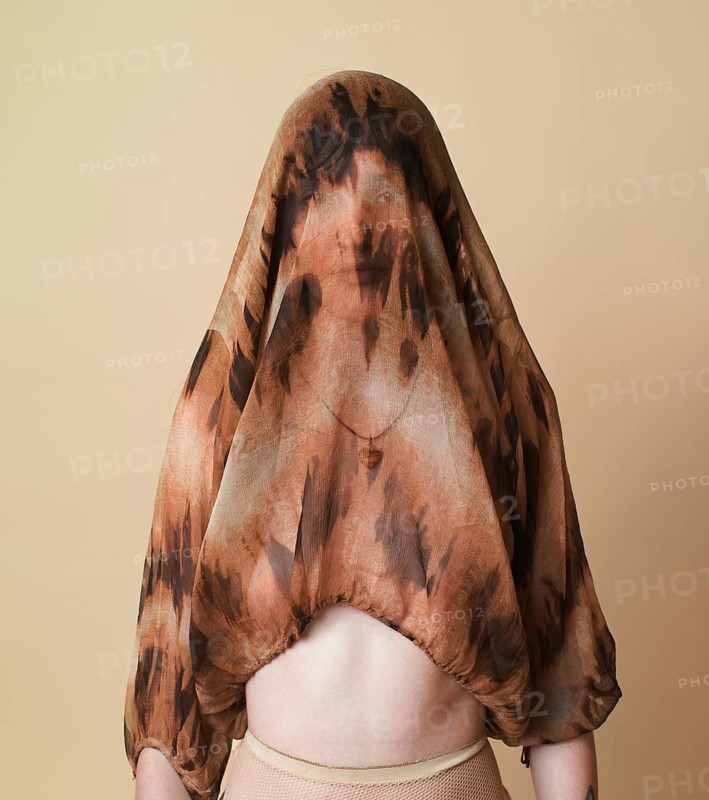 A young woman stands against a beige studio background, covering her head with a sheer, patterned top while staring directly at the camera