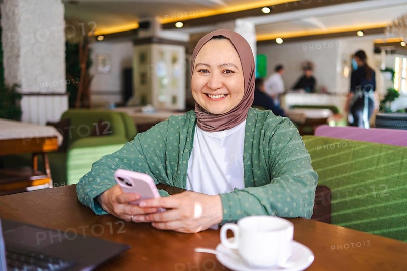Smiling Muslim woman in a green hijab using a smartphone in a cozy cafe. Warm ambiance with a laptop and cup of coffee on the table, highlighting casual modern lifestyle