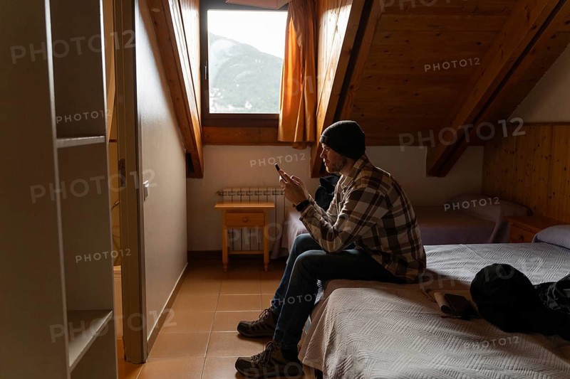 A man sits on a bed in a cozy cabin, engrossed in his smartphone. The room has a rustic wooden interior, enhanced by natural light from a window showing mountain views. He is dressed for winter in a beanie and plaid shirt