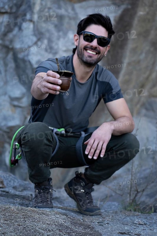 Vertical portrait of a smiling man squatting, wearing sunglasses and hiking gear, enjoys a traditional mate tea during a mountain adventure