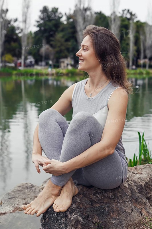 A young Caucasian woman with long brown hair sits on a rock by a calm lake. She wears a light gray outfit and smiles, surrounded by greenery and trees