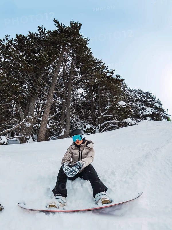 Young woman snowboarder wearing winter gear and goggles, sitting on a snowy mountain slope with tall pine trees under a clear sky, enjoying a break during winter sports