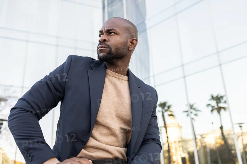 A confident business cuban man in a blazer poses outside a modern glass building. He looks determined, embodying professionalism and style in an urban business environment