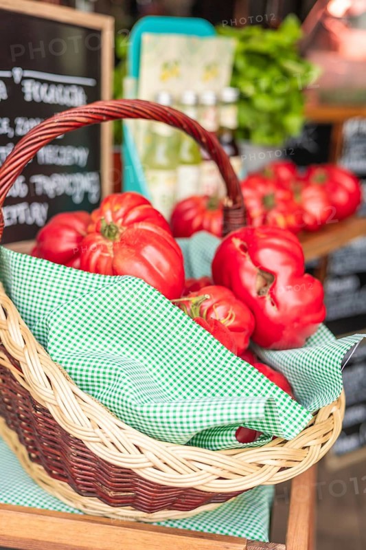 A wicker basket filled with ripe red tomatoes sits on a wooden table. The background features green leaves and a chalkboard with handwritten text