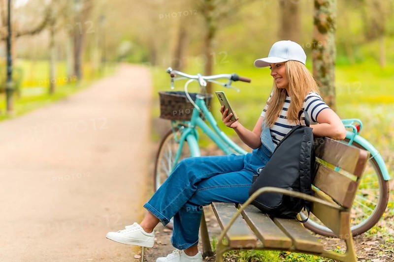 Cheerful young woman using smartphone while sitting on a bench in a park with her bicycle next to her, enjoying a sunny day outdoors