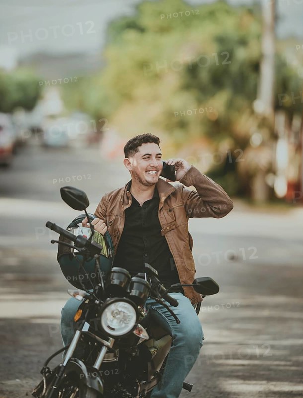 Handsome man sitting on his motorcycle making a phone call outdoors. Smiling young motorcyclist in jacket talking on cell phone