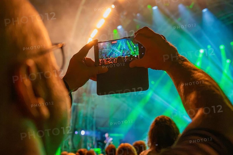 Person holding a smartphone, taking pictures of the concert with green stage lights and a crowd of people in the foreground, Calwer Klostersommer Festival, Hirsau Abbey, Calw, Germany