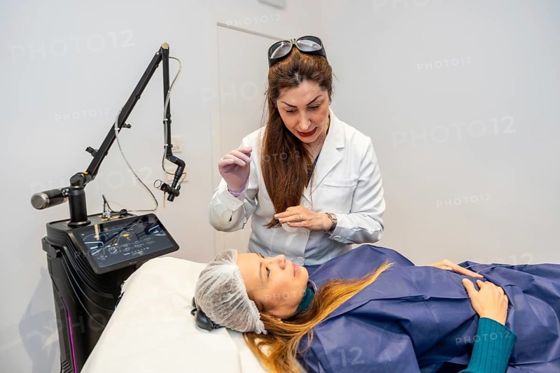 Doctor wearing a lab coat and safety glasses on her head cleaning a patient's face before a cosmetic treatment in an aesthetic clinic using modern laser equipment