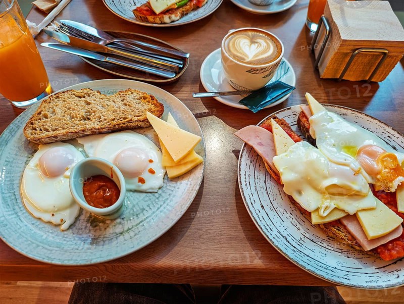 Close-up overhead view of a hearty breakfast spread on a wooden cafe table, featuring plates with fried eggs, toast, cheese, ham, coffee with latte art, and orange juice, ham sandwich