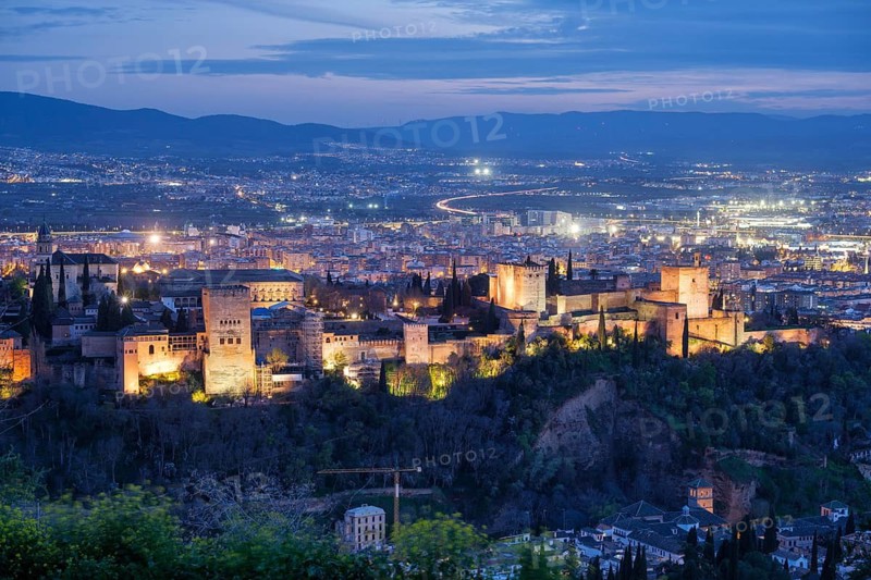 Elevated night view of the illuminated Alhambra fortress and palace complex in Granada, Spain, with the sprawling city lights and distant mountains during blue hour
