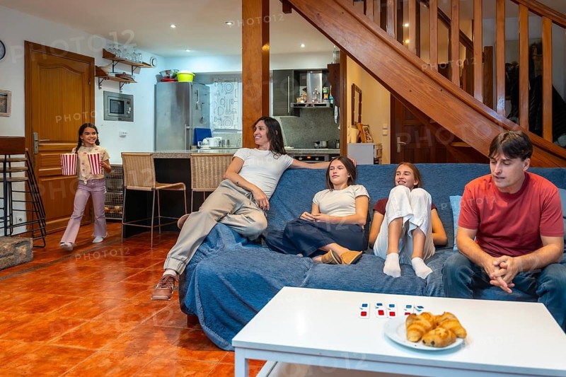 Family relaxing on a blue sofa in a cozy living room, watching tv together while a young girl brings popcorn and snacks for a warm evening of bonding and leisure