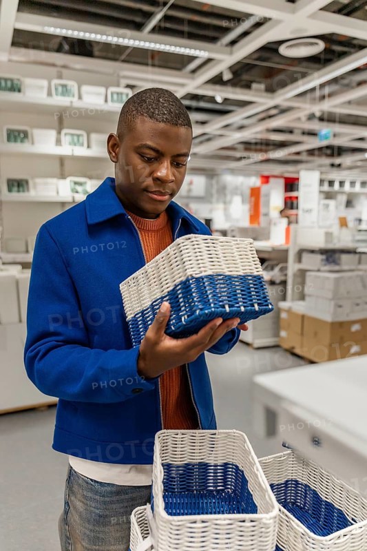 A young Black man examines a blue and white storage basket in a modern retail store. Shelves filled with various home goods are visible in the background