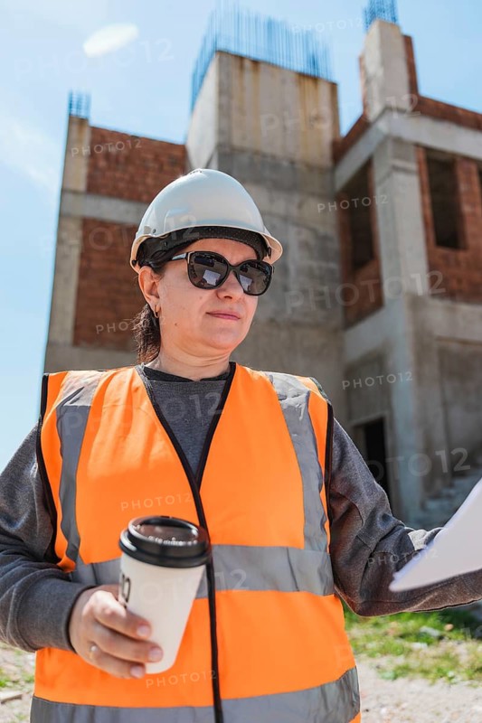 Woman in a hard hat and safety vest holds a coffee cup and construction plans at a building site. The background shows a partially constructed structure
