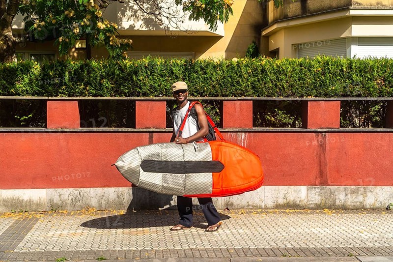 Happy young man walking along a sunny urban street carrying a surfboard in a protective bag, ready for a surfing adventure